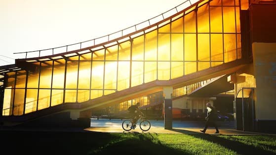 Golden hour view of a curved glass pedestrian bridge with a cyclist passing below